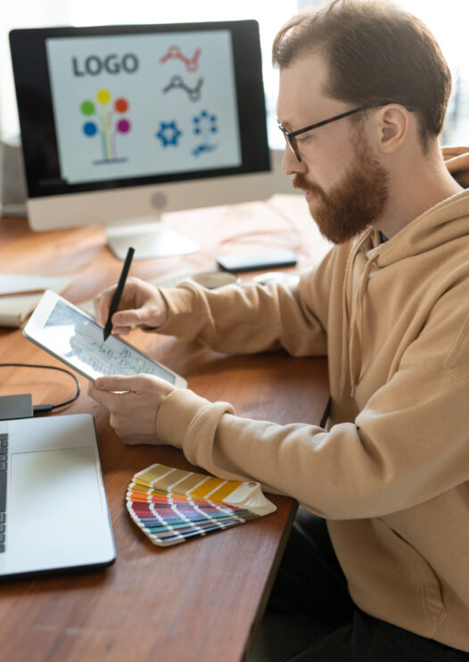 Concentrated bearded man in hoodie sitting at wooden table and using tablet while working on brand design